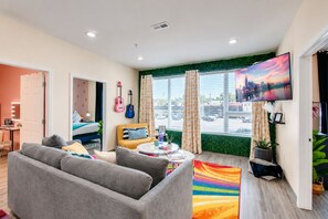 Una sala de estar moderna con un sofá gris, un sillón amarillo y una alfombra colorida. Hay guitarras en la pared, un televisor y una ventana con cortinas.