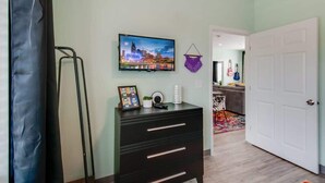 A room with a flat-screen TV mounted on the wall, a black dresser with drawers, a framed photo, and a hanging macrame wall hanging.