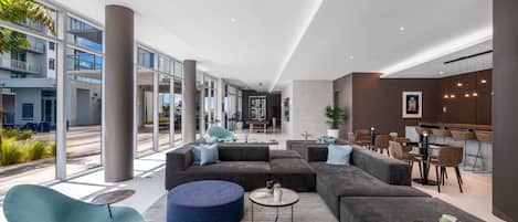 A modern lobby with a large gray sectional sofa, a teal chair, and a round ottoman. There is a dining area with wooden tables and chairs, and a large window offering a view of the building's exterior.