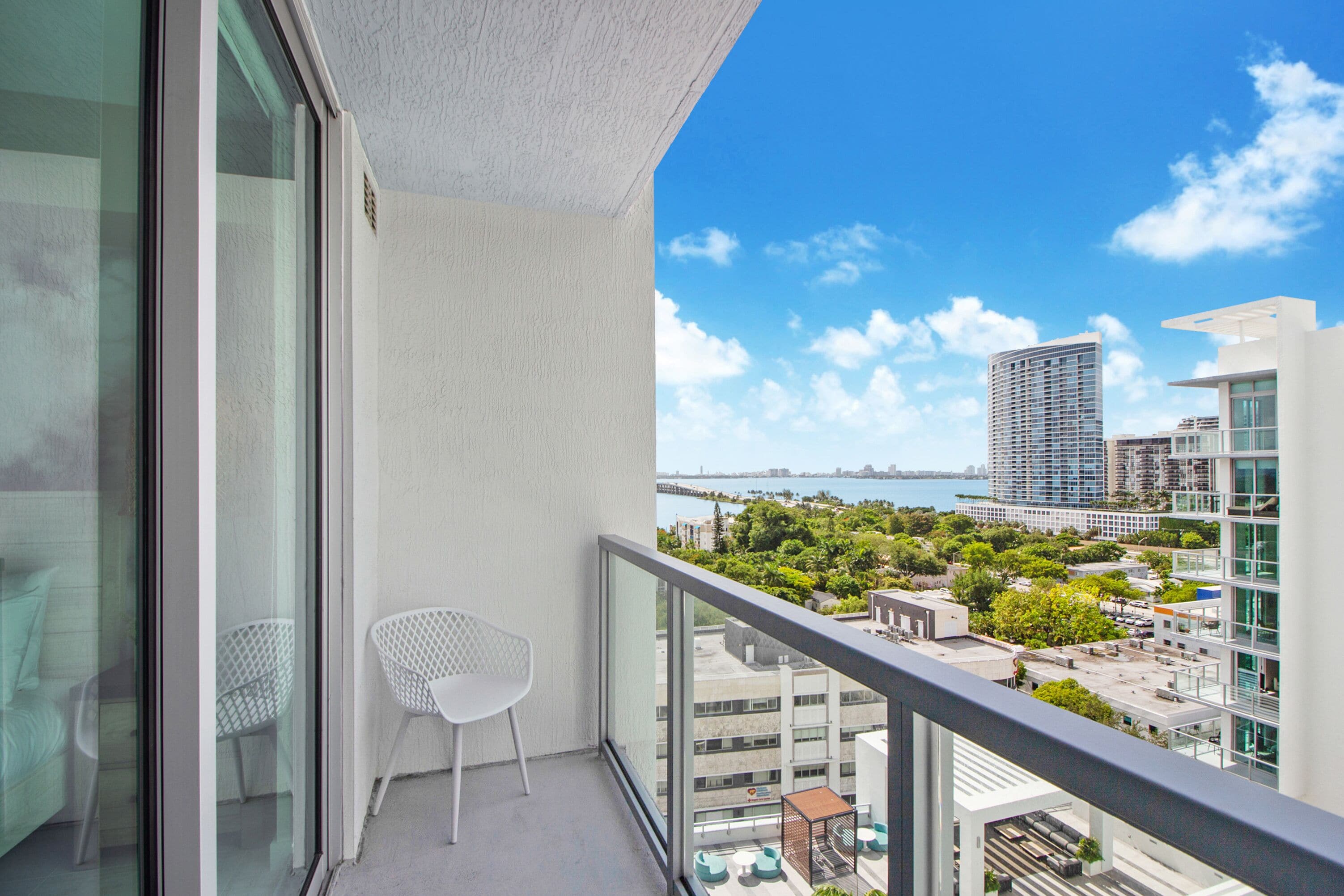 A balcony with a white chair, a glass door, and a view of a cityscape and water.