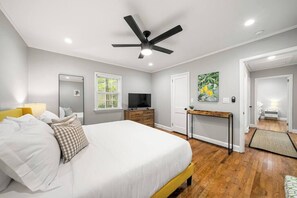 Une chambre à coucher comprenant un lit, un ventilateur de plafond, une télévision, un bureau et une porte donnant sur une autre pièce.