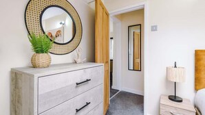 Une chambre avec une commode en bois, un miroir rond doté d’un cadre tressé, une lampe de chevet et une porte donnant sur une autre pièce.