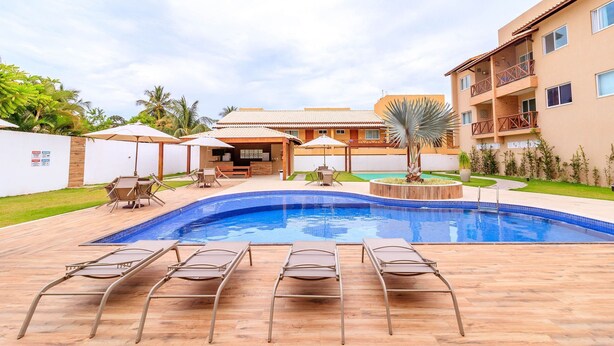 A swimming pool with lounge chairs and umbrellas, surrounded by a building with balconies and a garden. - GB37 Excellent Apt 2 Suites in Guarajuba (Camaçari)