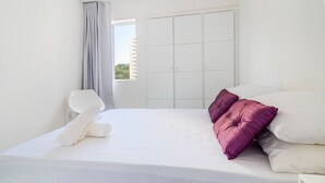A neatly made bed with white bedding and two purple pillows, a white bedside table, and a window with a view of buildings. - BR03 2 Bedroom Apt 200m from Porto Barra Beach (Salvador)