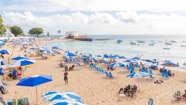 A beach with numerous blue umbrellas, sandy shore, and a few boats in the water. - BR03 2 Bedroom Apt 200m from Porto Barra Beach (Salvador)