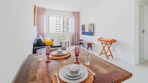 A modern living room with a wooden dining table set for four, a sofa, a TV, and a window with curtains. - BR03 2 Bedroom Apt 200m from Porto Barra Beach (Salvador)