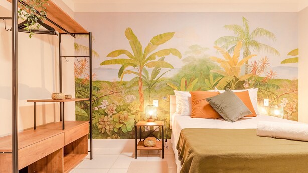 A bedroom with a bed, bedside tables, a wooden bench, a mural of tropical foliage, and a nightstand with a lamp. - VT06 Bedroom/Living Room Apartment - Vilas do Atlântico (Lauro de Freitas)