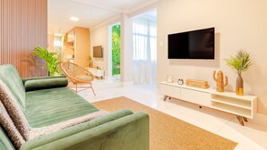 A modern living room with a green sofa, a flat-screen TV, a white console table, and a large window with a view of greenery. - VT06 Bedroom/Living Room Apartment - Vilas do Atlântico (Lauro de Freitas)