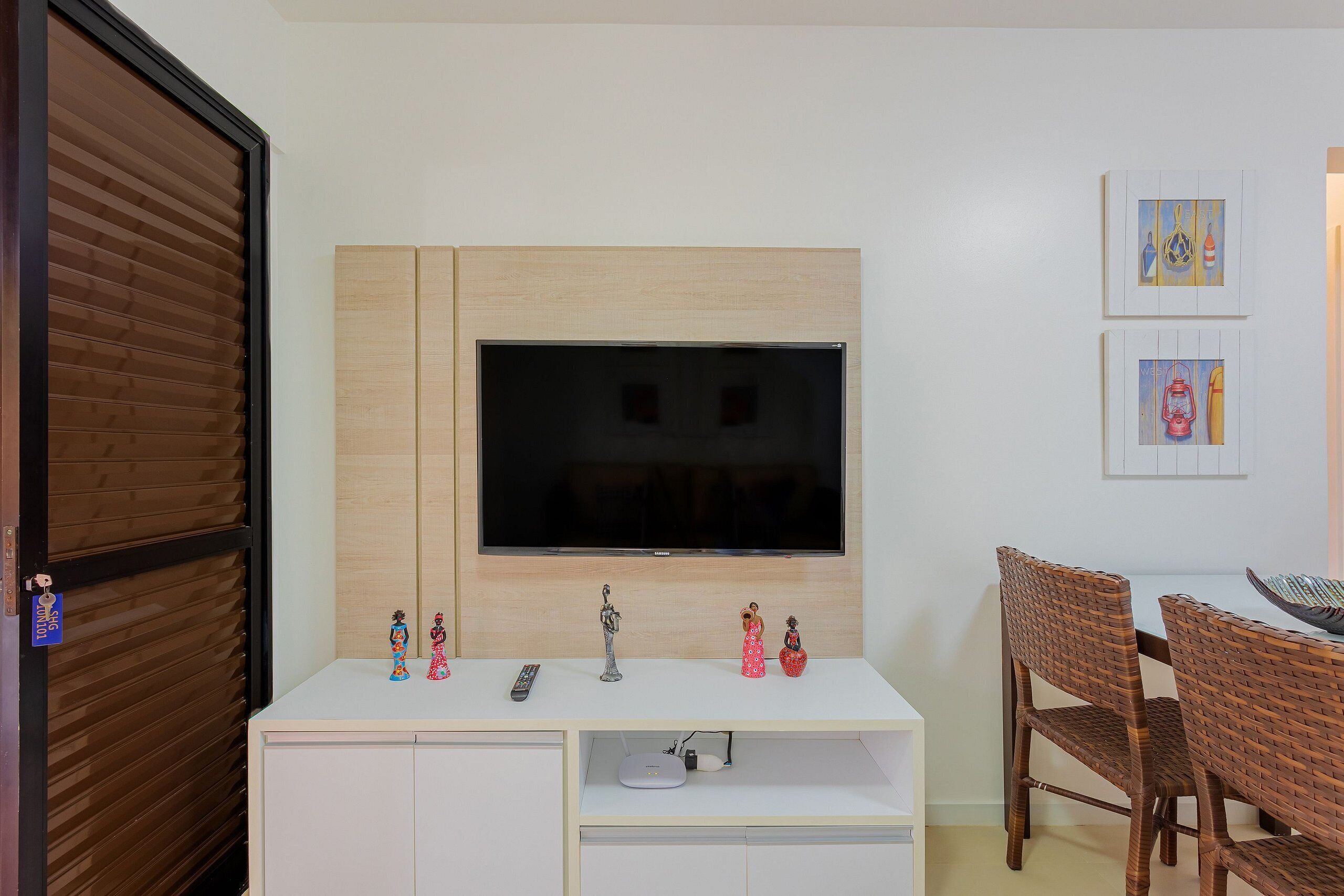Une chambre moderne avec un téléviseur fixé sur un panneau en bois, un meuble blanc en dessous, et un coin repas avec des chaises en osier.