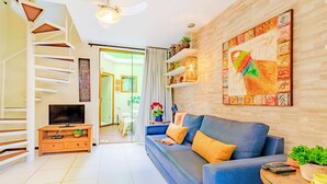 A modern living room with a blue sofa, a TV on a wooden stand, and a staircase with white railings. - IT26 Sea View Bungalow with 3 Suites, Right on the Sand - Varandas do Mar Residential (Camaçari)