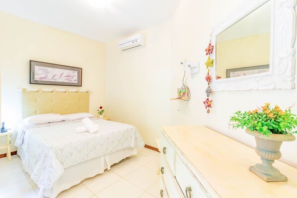 A bedroom with a bed, bedside table, dresser, mirror, wall-mounted air conditioner, and a potted plant.