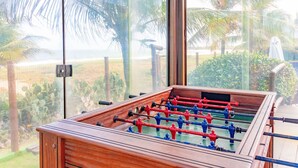 A foosball table with a view of a beach and palm trees through large windows. - IT26 Sea View Bungalow with 3 Suites, Right on the Sand - Varandas do Mar Residential (Camaçari)