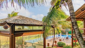 A glass-enclosed room with a view of a pool area, outdoor seating, and a thatched-roof structure. - IT26 Sea View Bungalow with 3 Suites, Right on the Sand - Varandas do Mar Residential (Camaçari)