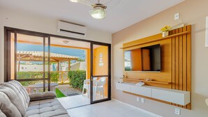 Un salon moderne avec un canapé, un téléviseur à écran plat fixé à un mur en bois, et une porte-fenêtre coulissante donnant sur un espace extérieur équipé d’une table et de chaises.