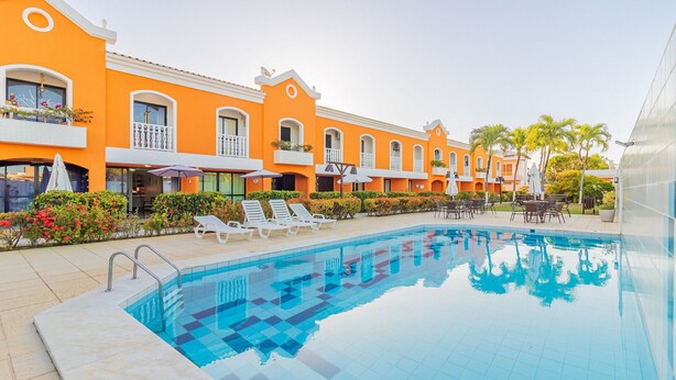 Une piscine entourée de chaises longues et de parasols, bordée de bâtiments aux façades orange, dotés de balcons et de fenêtres.