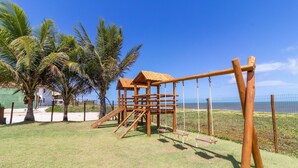 A wooden play structure with a roof, swings, and a slide, surrounded by grass and palm trees. - IT27 Village 3 Suites, Pé na Areia - Itacimirim (Camaçari)