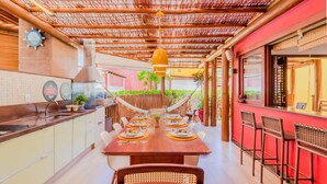 A restaurant with a long wooden table set for dining, a kitchen area with a stove, and a red wall with a window. - IT27 Village 3 Suites, Pé na Areia - Itacimirim (Camaçari)