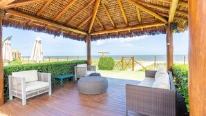 A covered outdoor seating area with wicker furniture, a wooden deck, and a view of the beach. - IT27 Village 3 Suites, Pé na Areia - Itacimirim (Camaçari)