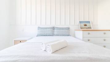 Una cama bien hecha con sábanas blancas y una toalla enrollada, una mesita de noche con una lámpara y un cuadro en un aparador.
