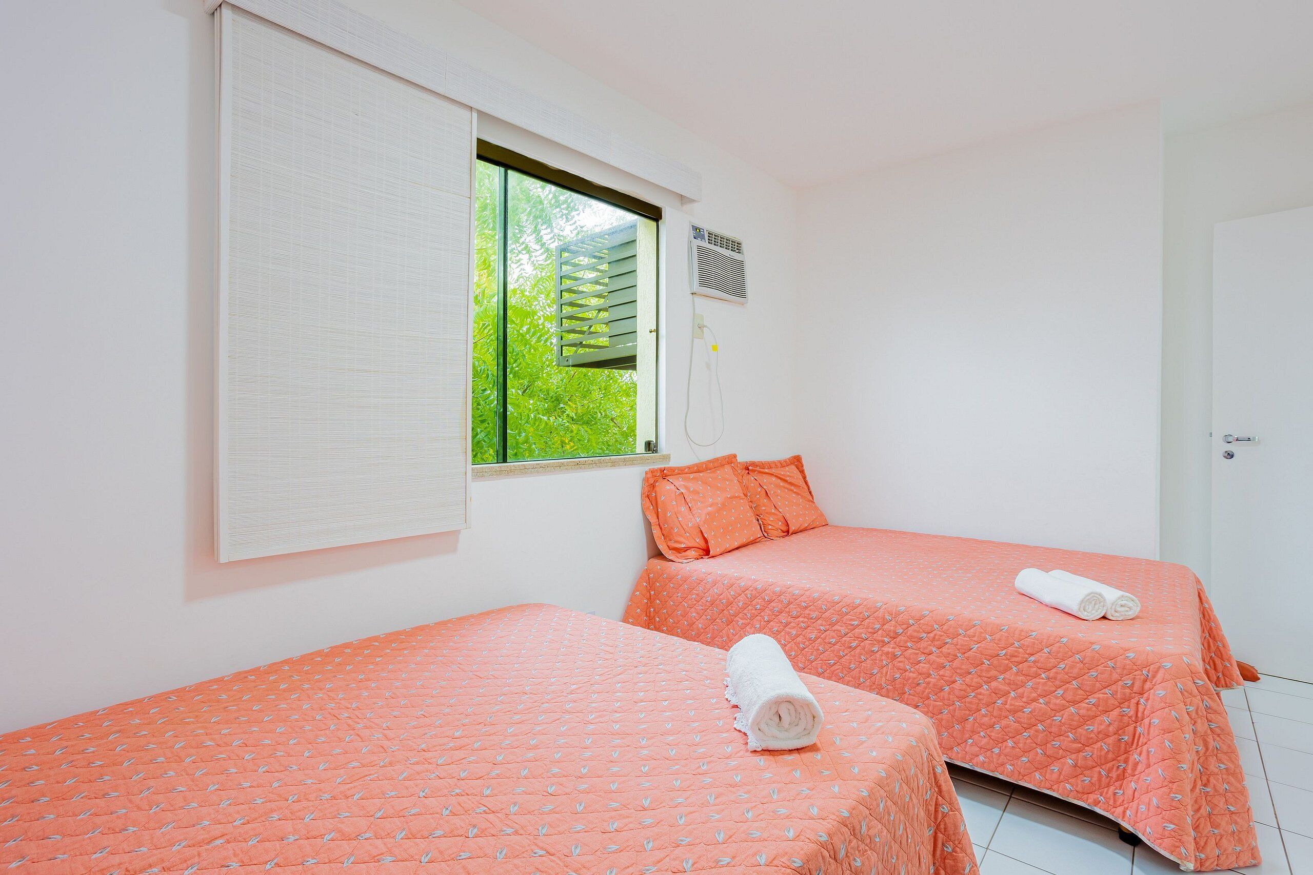 A room with two beds, a window with blinds, an air conditioning unit, and a door.