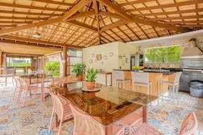 A covered outdoor dining area with a glass table and wicker chairs, surrounded by potted plants and a tiled floor.