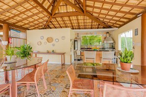 Un'area pranzo all'aperto coperta con tavolo in vetro, sedie in vimini e una cucina sullo sfondo.