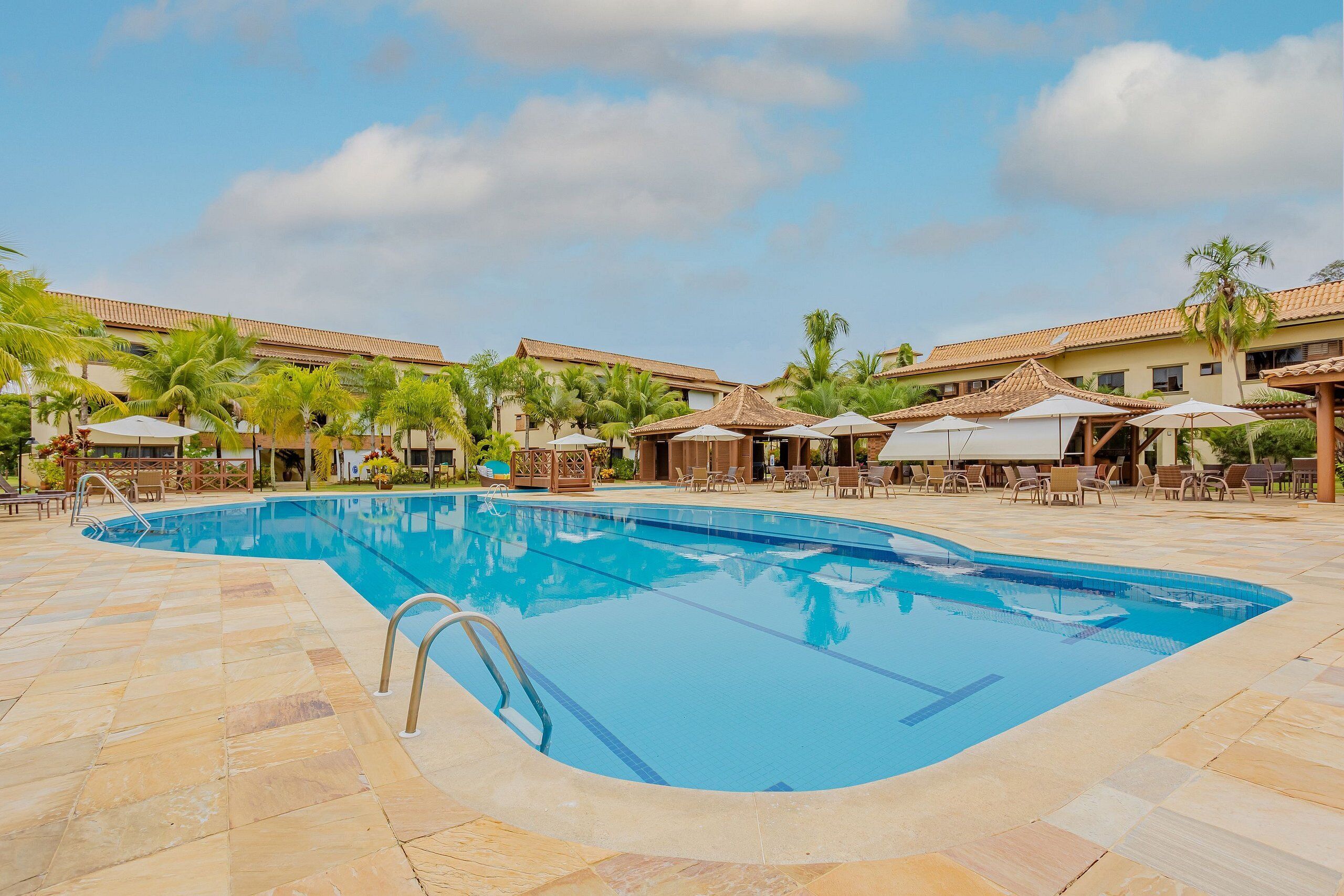 Piscina com deck revestido, rodeada por espreguiçadeiras e guarda-sóis, com palmeiras e edifícios ao fundo.