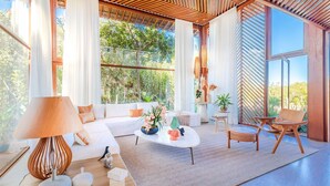 Ein modernes Wohnzimmer mit Sofa, Couchtisch und Sesseln, großen Fenstern mit Vorhängen und einer Holzbalkendecke.