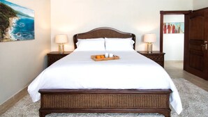A neatly made bed with white linens and a tray of oranges and cups, flanked by two nightstands with lamps, in a room with a wooden headboard and a painting of a coastal scene. - Porto Cupecoy- 3 Bedroom- Luxury Marina Penthouse (Sint Maarten)