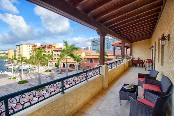 A balcony with wicker furniture, a view of a beach, and buildings in the background.