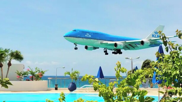 A large blue airplane with "KLM" written on it, landing at an airport near a pool area with palm trees and umbrellas. - Maho 1 bedroom condo beautifully decorated. (Sint Maarten)