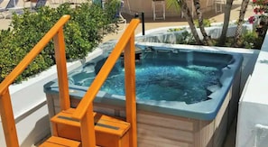 A hot tub with a wooden deck and orange railings, surrounded by greenery and a pool area.