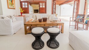 Een moderne woonkamer met een witte bank, een houten salontafel, twee zwarte geweven krukken, en een houten stoel met kussens.