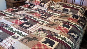 A bed with a quilt featuring animal prints, a wooden headboard, a nightstand with a lamp, and wall art. - A & B TRAIL HOUSE @ Hatfield-McCoy Trails, 5 Beds Plus Futon 2 Couches & 2 Baths (DELBARTON)