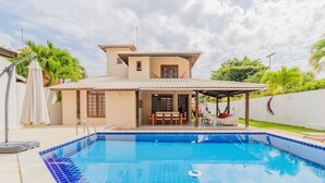 A swimming pool with a tiled edge, a covered patio area, and a house with a tiled roof and a door.