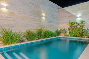 A swimming pool with a tiled edge, surrounded by plants and a light-colored wall with a built-in planter.