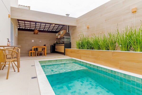 A swimming pool with tiled edges, a dining area with wicker furniture, and a kitchen area with a chimney.