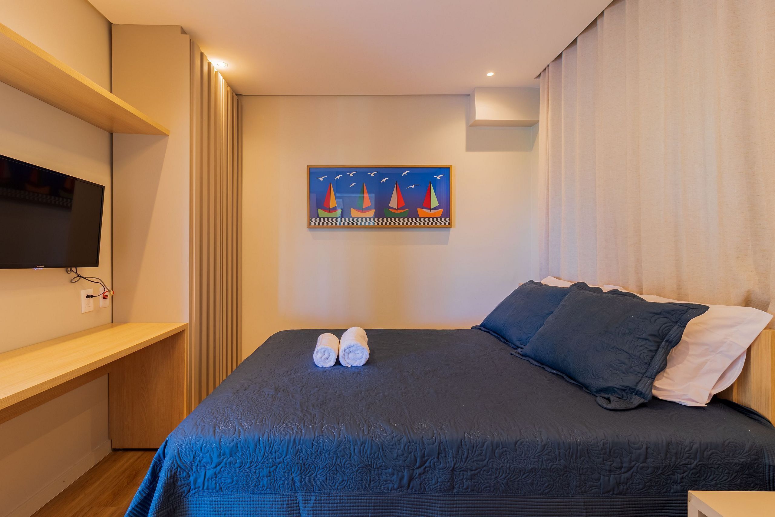 A neatly made bed with a dark blue bedspread and two rolled towels, a flat-screen TV mounted on a wooden shelf, and a painting of sailboats on the wall.