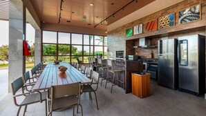 A modern kitchen with stainless steel appliances, a long dining table with chairs, and large windows offering a view of greenery.