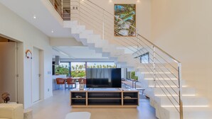 A modern living room with a staircase, a flat-screen TV, and a dining area with outdoor views.