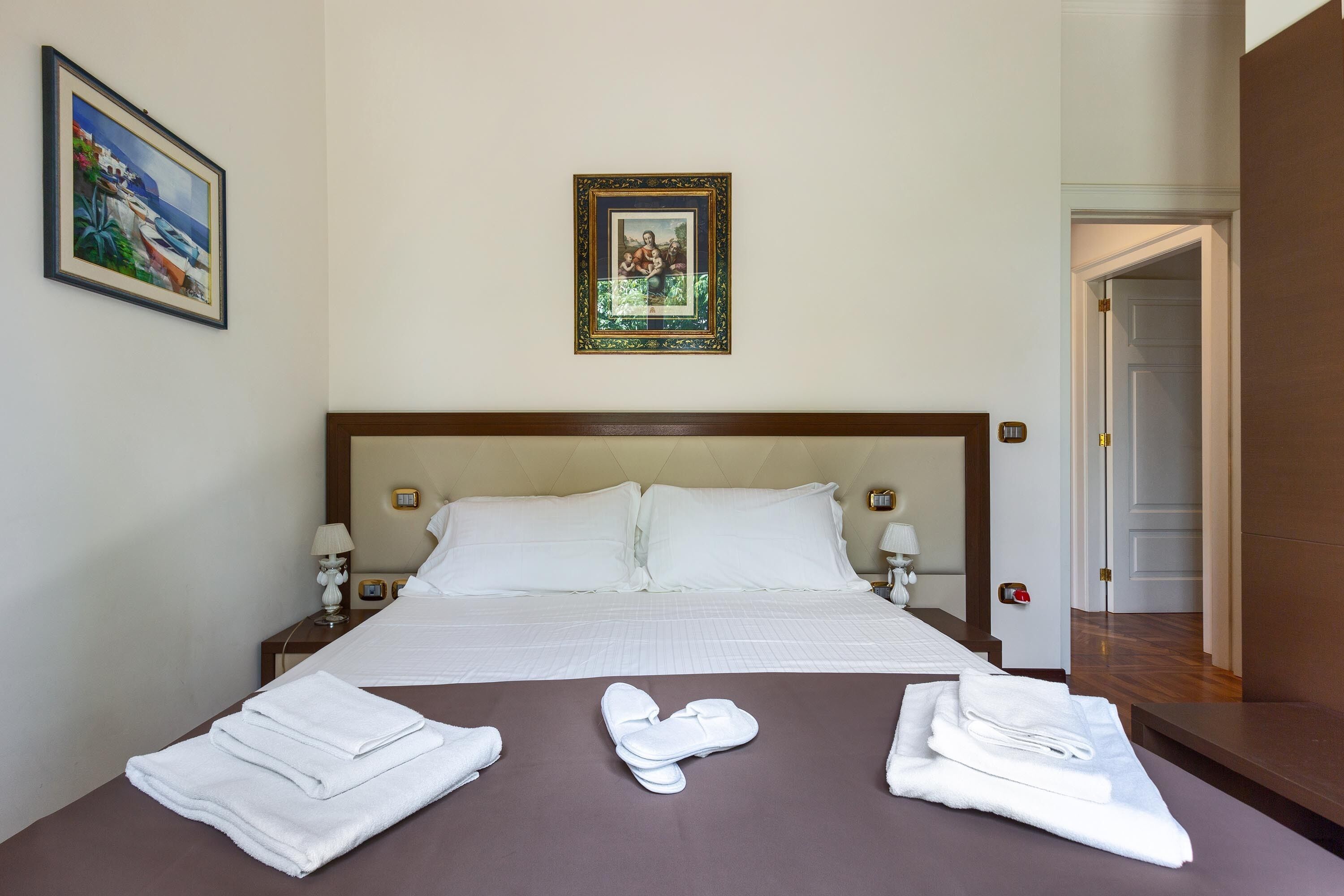 Una habitación de hotel con una cama grande, dos mesitas de noche con lámparas, un cuadro enmarcado en la pared y una puerta que da a otra habitación.