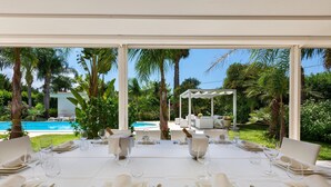 Une table dressée pour un repas, avec une nappe blanche, des couverts en argent et des verres, donnant sur une piscine et une végétation luxuriante.