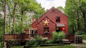 A red wooden cabin with a star decoration, surrounded by a wooden deck and lush greenery. - Mt Snow Chalet: Peaceful Escape w/Hot Tub (Wilmington)