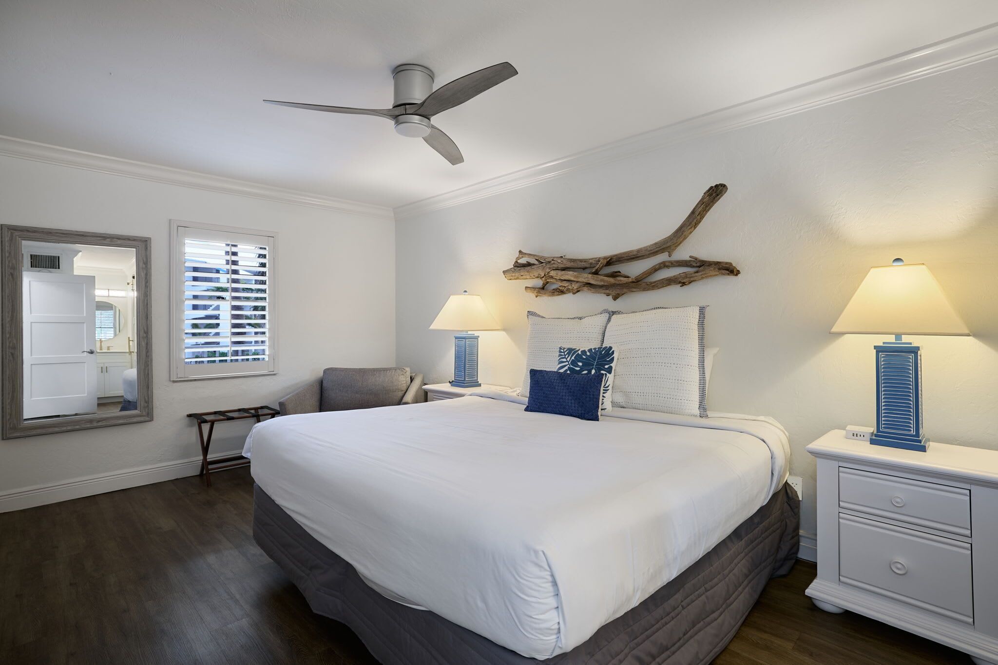 A bedroom with a large bed, two bedside tables with lamps, a ceiling fan, a mirror, and a window with blinds.
