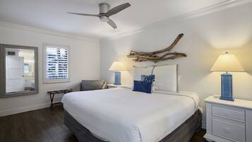 A bedroom with a large bed, two bedside tables with lamps, a ceiling fan, a mirror, and a window with blinds.
