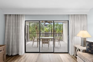 A room with a sliding glass door leading to an outdoor patio with a table and chairs, a lamp on a side table, and a wooden floor.