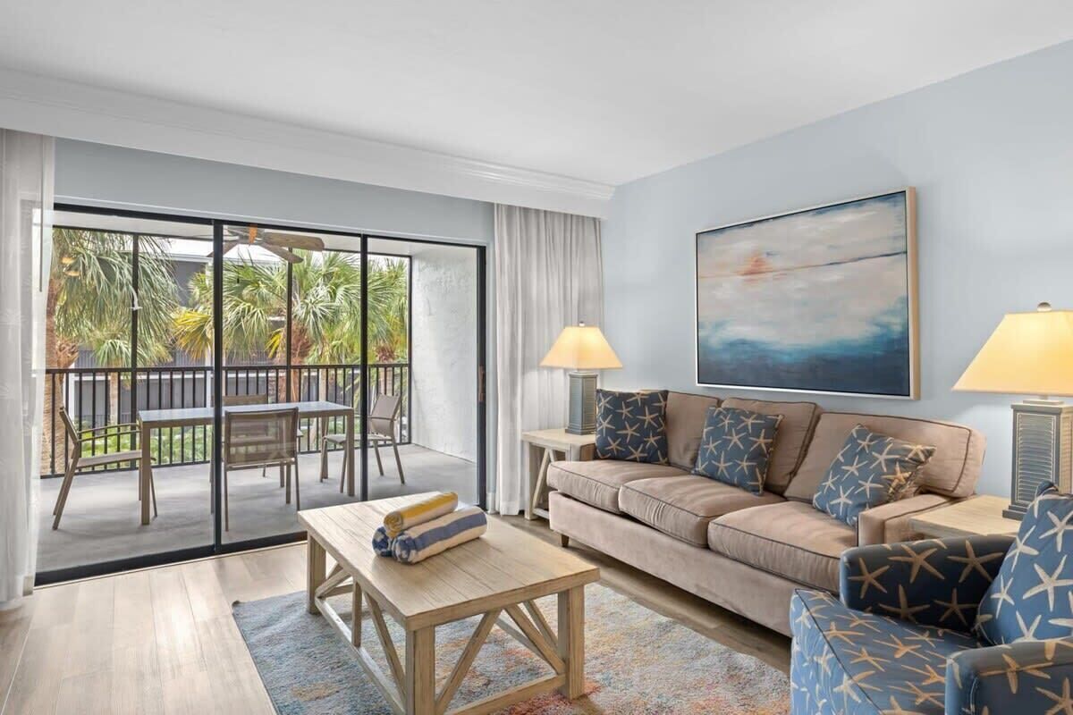 A living room with a sofa, coffee table, and chairs, a balcony with a table and chairs, and a painting on the wall.