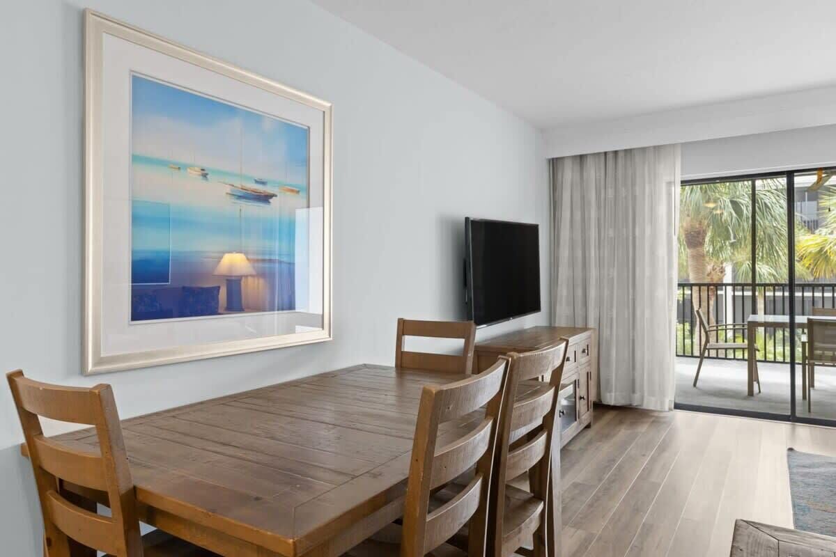 Une chambre moderne avec une table à manger en bois, des chaises, un téléviseur à écran plat et une grande fenêtre donnant sur des palmiers.