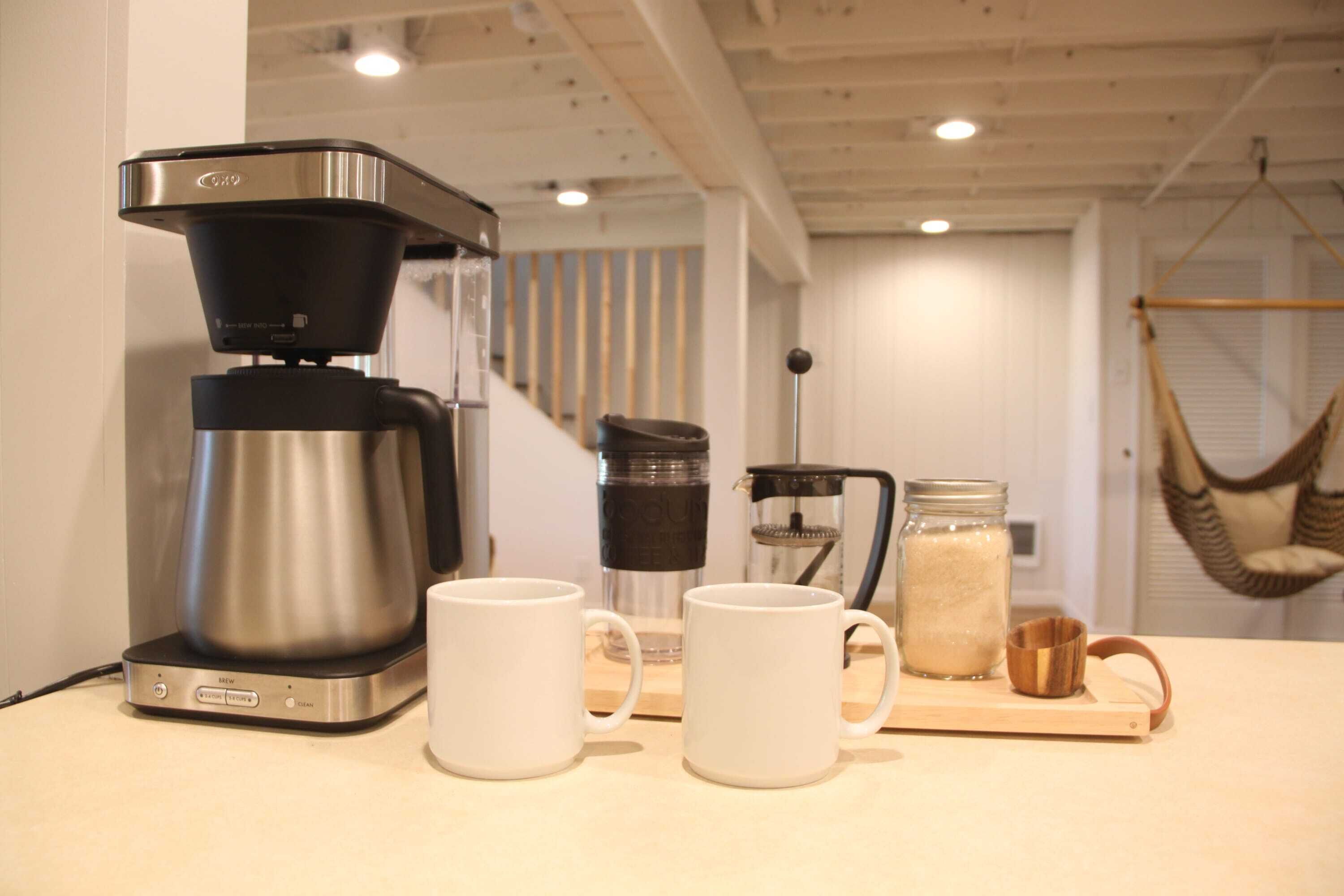 Une petite cuisine équipée d’une machine à café, de deux tasses blanches, d’un bocal en verre et d’une chaise suspendue.