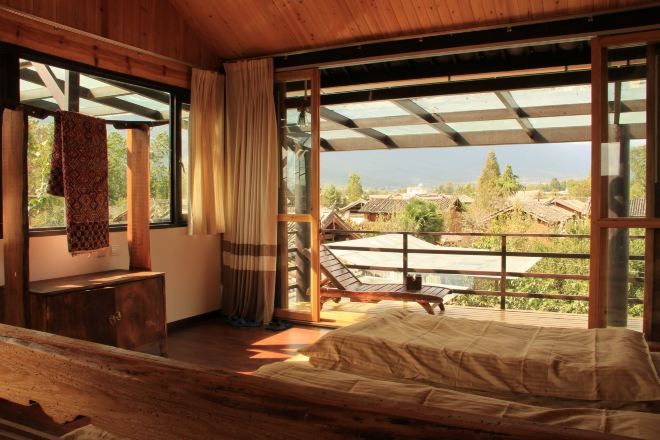 A wooden room with a bed, a bench, and a view of houses through large windows.